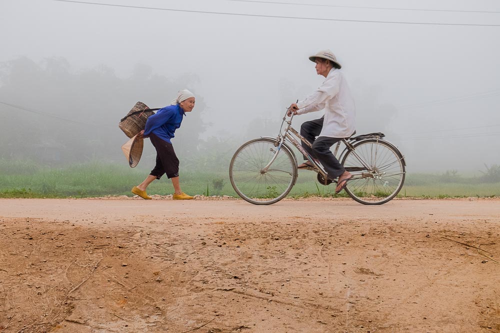 Tra le campagne e le piantagioni di riso una donna con una gerla e un uomo in bicicletta si incontrano lungo la strada
